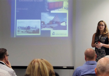 a lady with blond hair and glasses giving a presentation with a projection next to her 