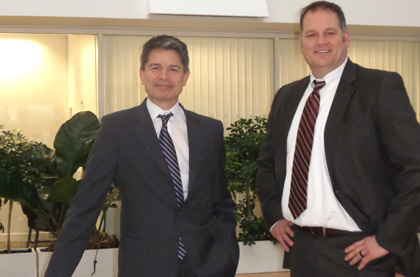 two men in dark suits, white shirts and stripy ties standing and smiling 