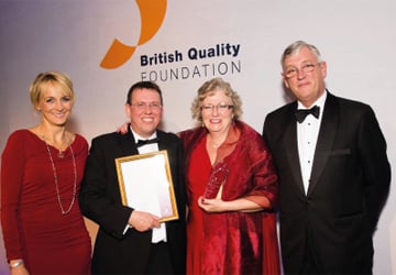 2 women in red dresses and 2 men in suits and black bow ties holding an award in front of a sign saying 'British Quality Foundation'
