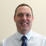 a headshot of a smiling man in a white shirt and blue tie in front of a cream wall