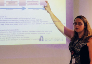 a lady with blonde hair and glasses pointing at a projection on a wall giving a presentation