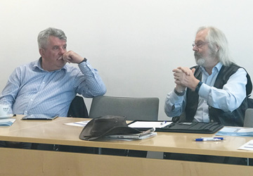 two men in blue shirts sat at a desk having a discussion