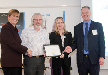 two women and two men in business dress shaking hands - the man and woman in the middle are holding a certificate