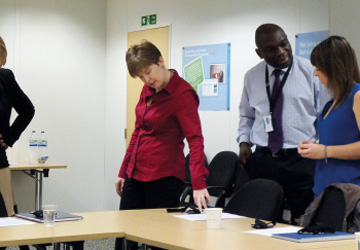 a woman in a red shirt looking a a paper on a desk next to a man in a shirt and tie talking to a woman in a blue dress