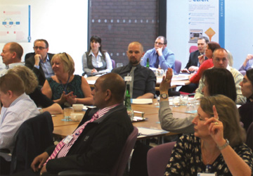 a room full of people sitting down at a desk, some of them have their hands up