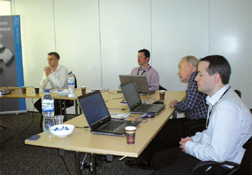 four men in business dress sitting at a desk with laptops open in front of them 