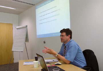 a man sat at a desk in a blue short sleeve shirt with a projection behind him