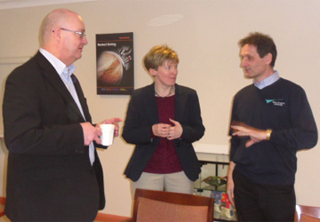 a man in a suit talking to a woman in a maroon top and a man in a shirt and jumper