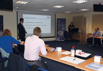 people in a business dress seated around a desk looking at a man pointing to a projector screen