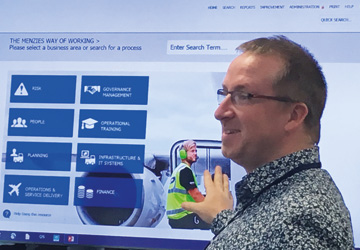 a close up of a man in a blue shirt with glasses pointing at a projection screen 