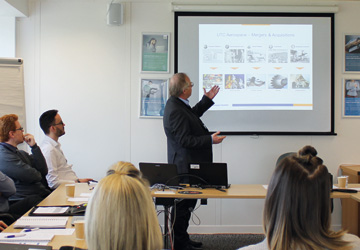 people in business dress sitting around a large desk looking at a man presenting in front of them