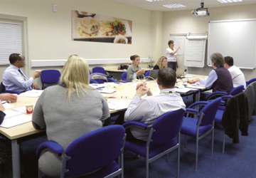 people in a business dress seated around a desk watching a man present 