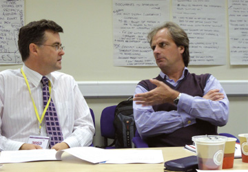 two men in business dress sat at a desk talking to each other 