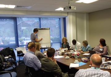 people in business dress sitting around a large desk looking at a woman presenting in front of them