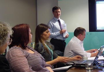 people sitting at a desk with a man in a shirt and tie standing behind them
