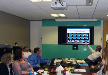 people in a business dress seated around a desk looking at a man pointing too a projector screen 