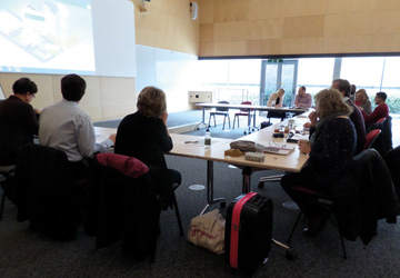 people in business dress sitting around a large desk looking at a woman presenting in front of them