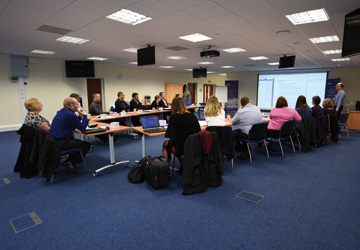 people sitting around a U shaped desk looking at a projection 