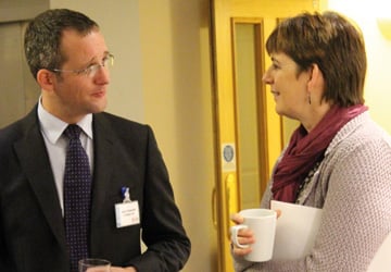 a man in a suit with a name tag and glasses talking to a woman in a pink cardigan holding a mug 