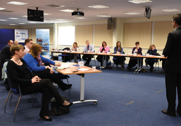 people in business dress sitting at two long desks opposite each other, looking at a man in a suit