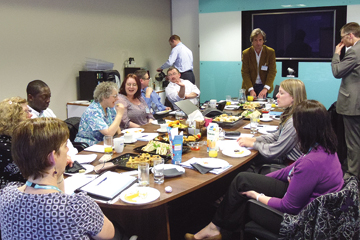 a room full of people in business dress some stood up some sat down around a desk with food on it, chatting to each other