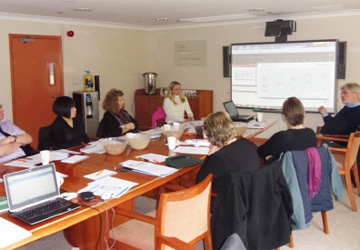 people sat along a long desk looking at a screen projector