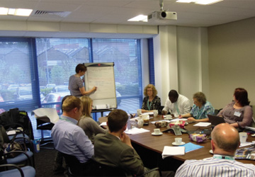 people in business dress sitting around a large desk looking at a woman presenting in front of them