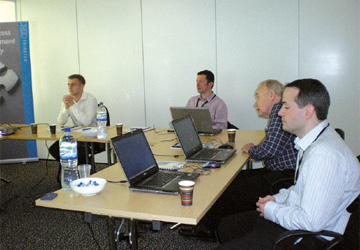 4 men at a desk with laptops in front of them