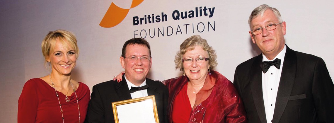 2 women in red dresses and 2 men in suits standing and smiling, one of the men is holding an award