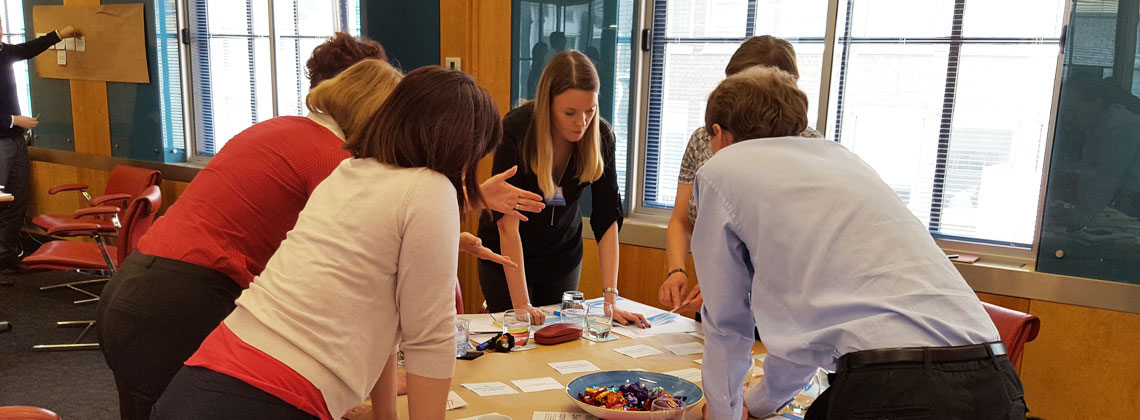 6 people standing around a table, leaning on it, looking at pieces of paper on the table 