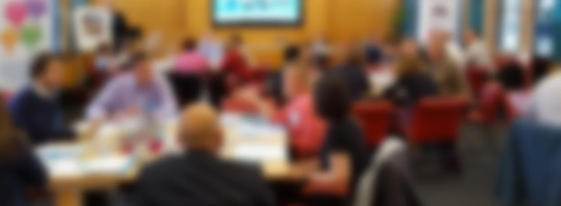 blurred photo of people sitting at round tables looking at a screen on a wall 