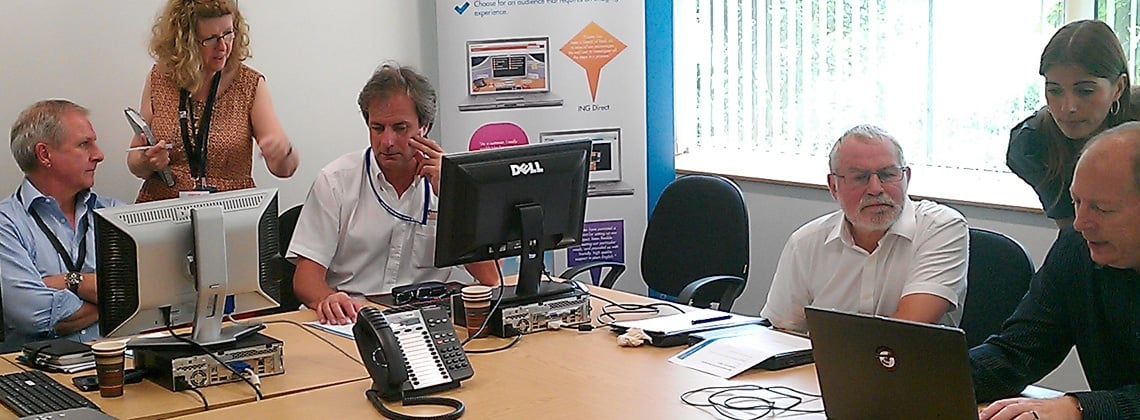 four men sitting at a desk on computers and two women looking at the screens helping them